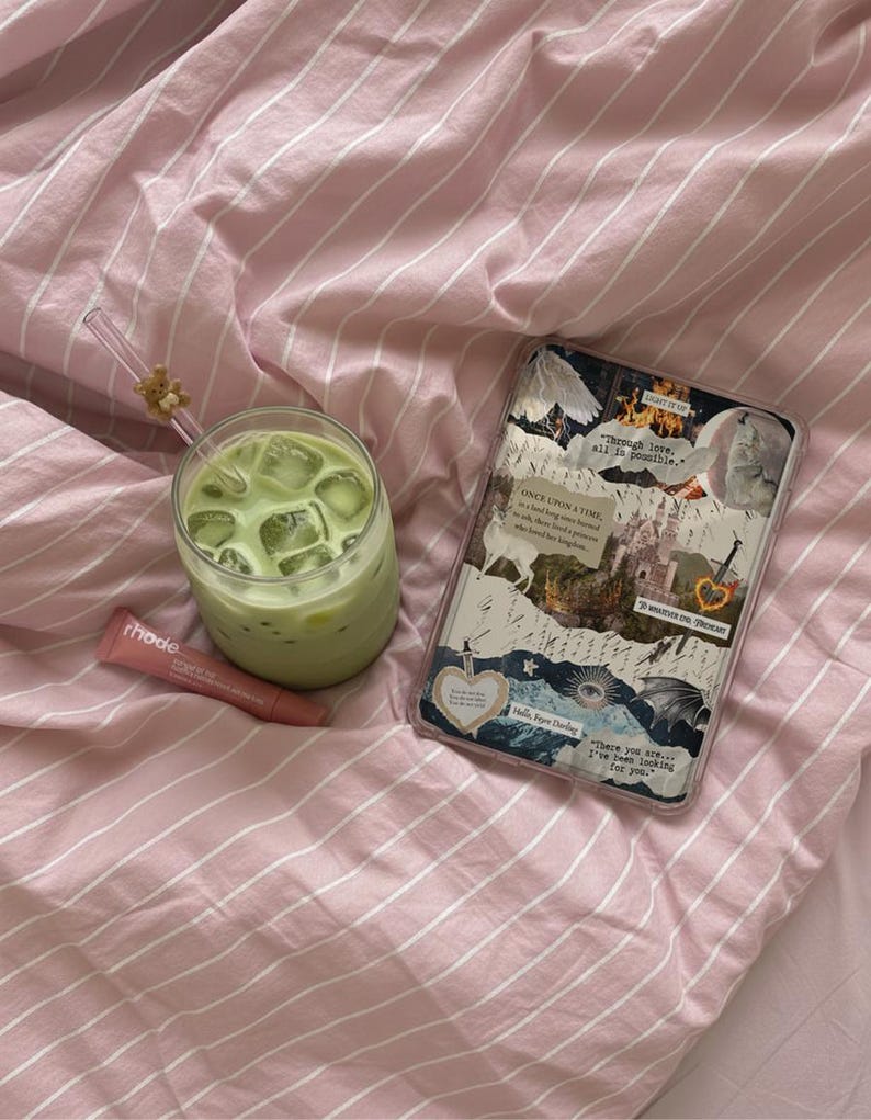 May include: A glass of iced matcha latte with a bear-topped straw, a pink Rhode lip treatment tube, and a rectangular tablet case featuring a collage of fantasy-themed imagery and text including "Through love, all is possible" and "Hello, Feyre Darling."