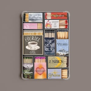 Könnte beinhalten: Ein rechteckiges Tablett mit zwölf verschiedenen Streichholzschachteln mit unterschiedlichen Designs und Texten, darunter The World Will Be Saved & Remade By The Dreamers, Light It Up, Suriel Tea, The Night Court, Velaris, Lunathion, Terrasen, Fireheart und Starborn.