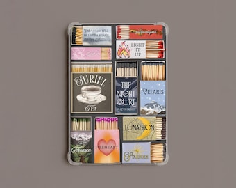 May include: A rectangular tray containing twelve assorted matchboxes with various designs and text including The World Will Be Saved & Remade By The Dreamers, Light It Up, Suriel Tea, The Night Court, Velaris, Lunathion, Terrasen, Fireheart, and Starborn.