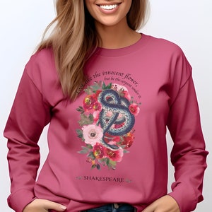 Op de afbeelding: Een roze sweatshirt met een bloemenprint en een slang. De tekst "Look like the innocent flower, but be the serpent under it" staat op het sweatshirt gedrukt. De tekst "SHAKESPEARE" staat onder de print.