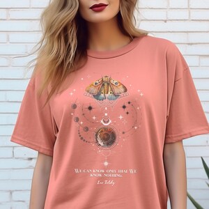 May include: A coral coloured t-shirt with a graphic print of a moth, celestial bodies, and stars. The text "We can know only that we know nothing. Leo Tolstoy" is printed on the shirt.