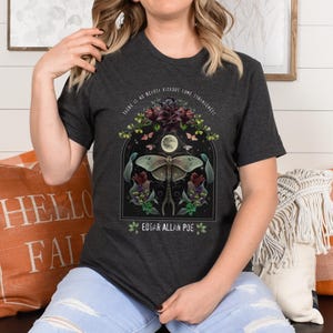May include: Dark grey t-shirt with a graphic print of a moth, flowers, and a crescent moon. The text "There is no beauty without some strangeness" and "Edgar Allan Poe" are printed on the shirt.