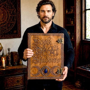 Puede incluir: Un libro encuadernado en cuero marr&oacute;n con el t&iacute;tulo "Book of Shadows" en relieve en la portada. La portada presenta un pentagrama, lunas crecientes y una piedra preciosa azul. El libro tiene cierres met&aacute;licos y bordes decorativos.