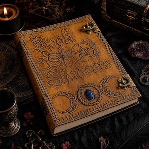 Puede incluir: Un libro encuadernado en cuero marr&oacute;n con las palabras "Book of Shadows" grabadas en la portada. La portada presenta un pentagrama, medias lunas y una piedra azul. El libro tiene cierres de lat&oacute;n y bordes decorativos.