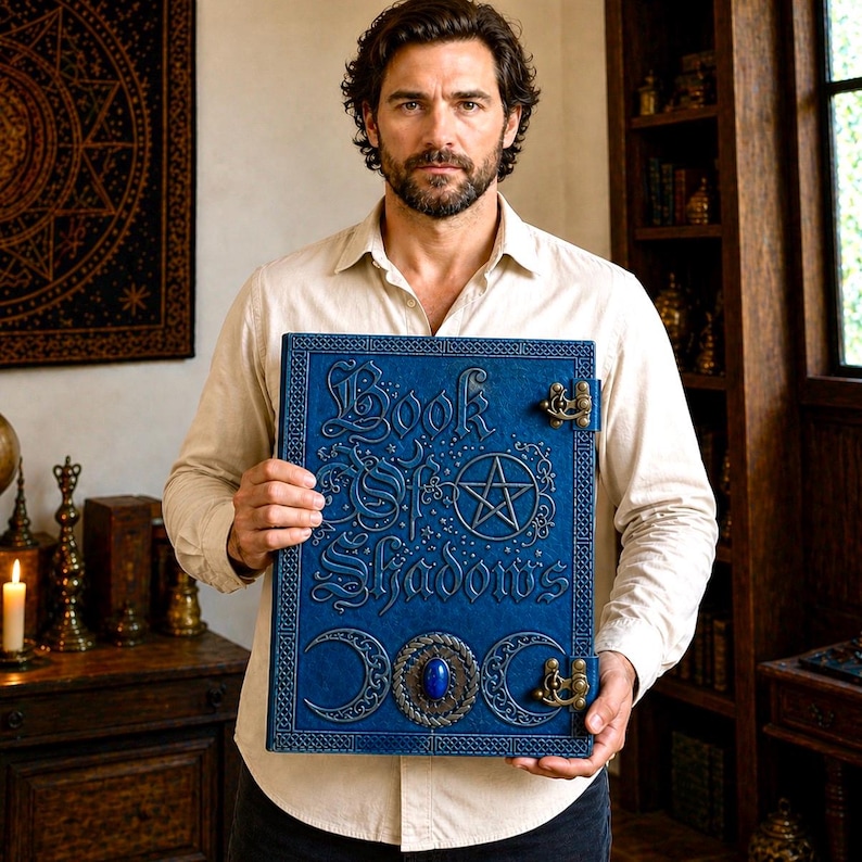 Puede incluir: Un libro azul encuadernado en cuero titulado "Book of Shadows" con letras doradas ornamentadas y un dise&ntilde;o de pentagrama. El libro presenta cierres decorativos y una gema azul central. El libro se sostiene frente a una camisa de color claro.