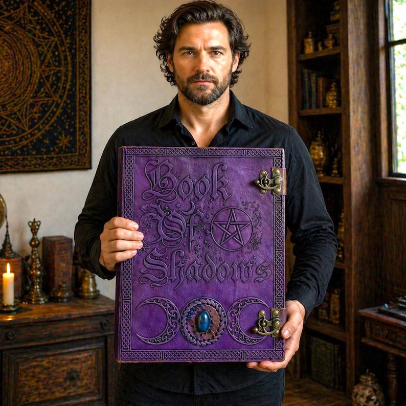 Puede incluir: Un libro grande de cuero p&uacute;rpura titulado "Book of Shadows" con letras en relieve y un dise&ntilde;o de pentagrama. El libro presenta cierres met&aacute;licos decorativos y una piedra preciosa azul. El libro es sostenido frente a una persona.