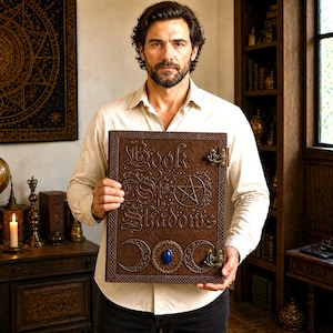 Puede incluir: Un libro encuadernado en cuero marr&oacute;n titulado "Book of Shadows" con letras en relieve y un dise&ntilde;o de pentagrama. El libro presenta una piedra preciosa azul y cierres decorativos. El libro est&aacute; sostenido frente a una persona.