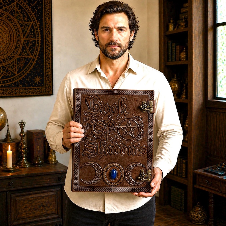 Puede incluir: Un libro encuadernado en cuero marr&oacute;n titulado "Book of Shadows" con letras en relieve y un dise&ntilde;o de pentagrama. El libro presenta una piedra preciosa azul y cierres decorativos. El libro est&aacute; sostenido frente a una persona.