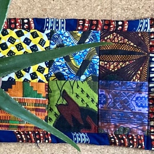 May include: A colourful patchwork table runner with a blue background and a variety of geometric patterns in shades of brown, yellow, green, and blue. A seashell and a small wooden box are placed on the runner, and a green succulent plant is in the foreground.