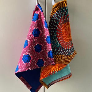 May include: Two hanging kitchen towels featuring vibrant patterns. One towel has a magenta background with blue floral motifs and a solid blue reverse. The other towel displays a geometric orange, yellow, and teal circular design with a textured light blue reverse.