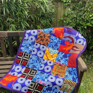 May include: A patchwork quilt with a purple border. The quilt is made of various colorful fabrics with geometric patterns. The quilt is folded and lying on a wooden bench.