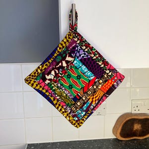 May include: A square quilted potholder featuring a vibrant patchwork of African wax print fabrics in yellow, green, red, blue, and purple. The textile accessory has a fabric loop at the top corner for hanging and displays a variety of geometric patterns.