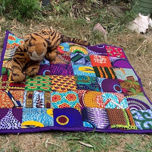 May include: A colorful patchwork quilt with a purple border, featuring various patterned fabric squares in vibrant hues. A stuffed tiger toy with orange and black stripes rests on top of the quilt, creating a playful scene.