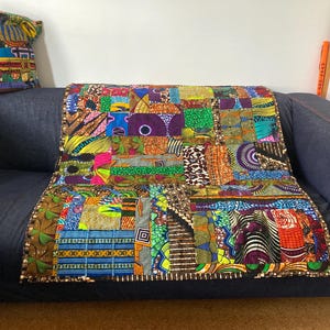 Peut inclure: Une courtepointe patchwork colorée et un coussin assorti reposent sur un canapé bleu foncé. La courtepointe présente une variété de carrés de tissu à motifs dans des tons de vert, de bleu, de jaune et de rouge. Le coussin a un design similaire. Le canapé est contre un mur blanc.