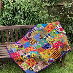 May include: A vibrant patchwork quilt with a purple border, displayed on a wooden bench. The quilt showcases a variety of colourful African fabric patterns in red, yellow, blue, and green. The quilt is placed outside on a sunny day.