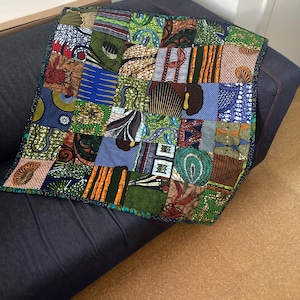 May include: A square patchwork quilt with a variety of colorful African fabric patterns. The quilt features a mix of geometric and floral designs in shades of blue, green, orange, and brown. The edges are finished with a dark blue border.