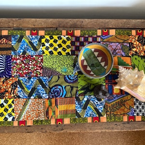May include: A colourful patchwork table runner featuring a green and brown ceramic bowl, a seashell, and a small wooden figure. The runner is made from various fabrics with geometric and floral designs.