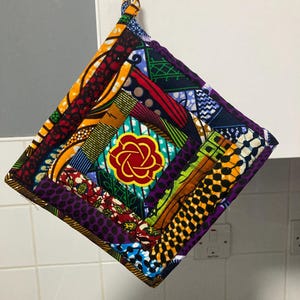Könnte beinhalten: Ein farbenfroher, gesteppter Topflappen mit einem lebendigen Patchwork-Design. Der quadratische Topflappen hat ein zentrales Blumenmotiv und besteht aus verschiedenen gemusterten Stoffen in Rot-, Gelb-, Blau- und Lilatönen. Er hat eine Schlaufe zum Aufhängen.