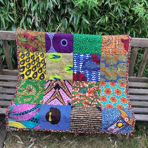 May include: A colorful patchwork quilt with a variety of African wax print fabrics. The quilt features squares of different patterns and colors, including yellow, green, purple, and orange. The edges are trimmed with a red beaded border.