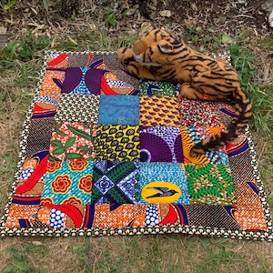 Puede incluir: Una colcha de retazos rectangular que presenta una variedad de vibrantes patrones de tela de inspiración africana en tonos naranja, azul, morado, verde y amarillo, con un tigre de peluche con rayas negras y vientre beige encima.