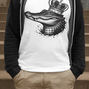 May include: A white t-shirt with a black and white graphic of a crocodile wearing a crown. The crocodile is facing to the right and has a detailed design.