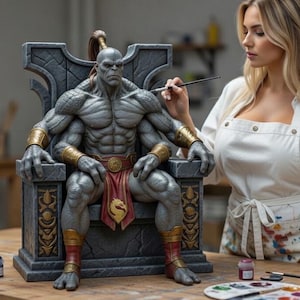 Pode incluir: Estátua detalhada de um personagem de quatro braços e pele cinzenta sentado em um trono. A figura tem braceletes dourados e um tapa-sexo vermelho. Uma pessoa está pintando a estátua com um pincel. Potes de tinta e uma paleta sobre a mesa.
