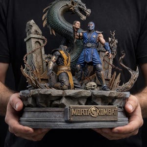 May include: A detailed statue featuring characters from Mortal Kombat. The figures include a blue-clad warrior, a kneeling figure in yellow, and a dragon. The base has the Mortal Kombat logo. The statue is held by a person.