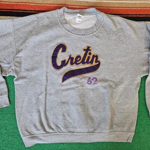 May include: A heather gray crewneck sweatshirt featuring the word Cretin in purple script with a gold outline across the chest. Below the text, the number 62 is embroidered in purple. The garment has long sleeves and a ribbed collar.