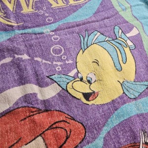 Op de afbeelding: Een paarse strandhanddoek met een grafische print van De Kleine Zeemeermin met gele tekst, een gele vis met blauwe vinnen en een roodharig personage. Het ontwerp bevat lichtblauwe golvende lijnen en witte bubbels op een paarse achtergrond.