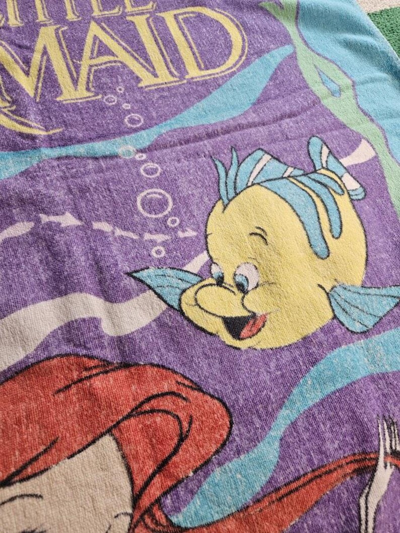 Op de afbeelding: Een paarse strandhanddoek met een grafische print van De Kleine Zeemeermin met gele tekst, een gele vis met blauwe vinnen en een roodharig personage. Het ontwerp bevat lichtblauwe golvende lijnen en witte bubbels op een paarse achtergrond.