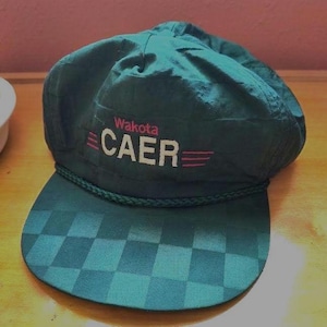 May include: A dark teal cap with the word "Wakota" above "CAER" in white text with pink accents. The brim features a checkered pattern. A braided rope trim accents the brim.