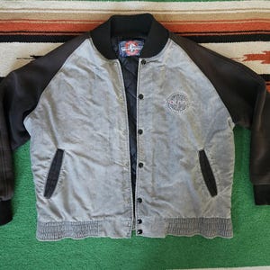 May include: A gray and black varsity-style jacket featuring long black sleeves, a black ribbed collar, and a black ribbed hem. The front has a snap-button closure, two side pockets, and a circular embroidered logo on the chest reading Polar.