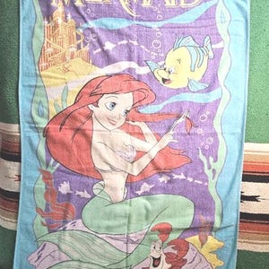 Op de afbeelding: Een rechthoekige strandhanddoek met een lichtblauwe rand en een centrale paarse afbeelding van Disney's De Kleine Zeemeermin. Het ontwerp bevat Ari&euml;l met rood haar, Botje, Sebastiaan de krab en een gouden onderwaterkasteel.