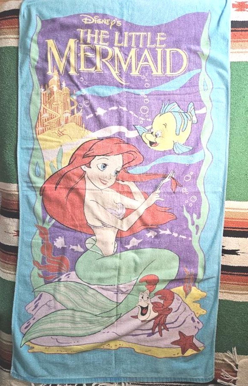 Op de afbeelding: Een rechthoekige strandhanddoek met een lichtblauwe rand en een centrale paarse afbeelding van Disney's De Kleine Zeemeermin. Het ontwerp bevat Ari&euml;l met rood haar, Botje, Sebastiaan de krab en een gouden onderwaterkasteel.