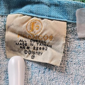 Op de afbeelding: Een lichtblauwe katoenen handdoek met een wit stoffen label met de tekst FRANCO, ALL COTTON, MADE IN TAIWAN, RN 22682 en Disney. Het label bevat een cirkelvormig logo met een gestileerde letter S en een copyright-symbool op de stof.