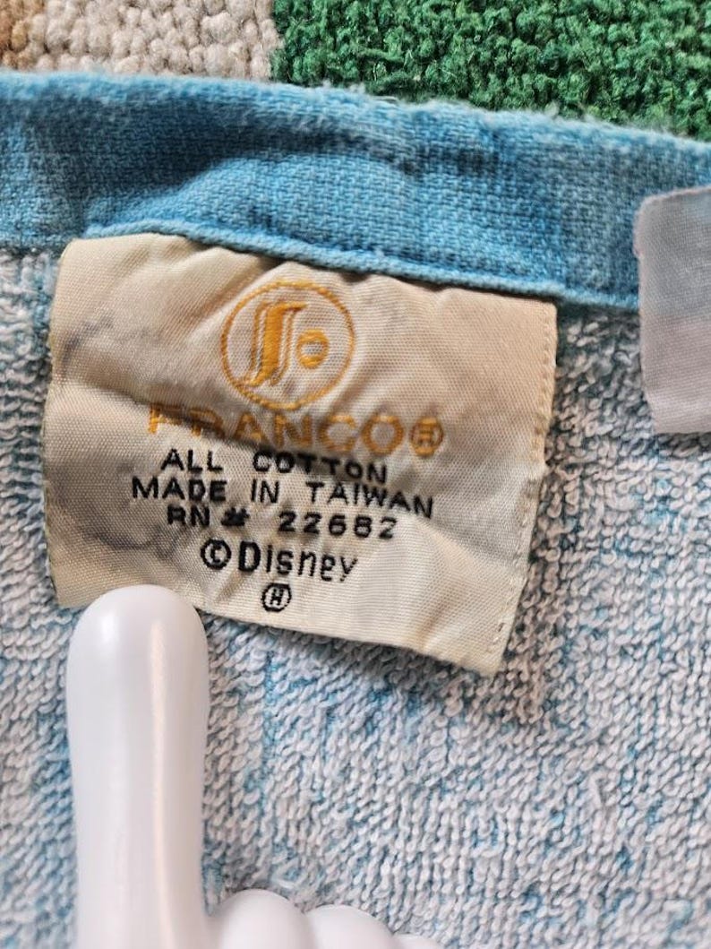 Op de afbeelding: Een lichtblauwe katoenen handdoek met een wit stoffen label met de tekst FRANCO, ALL COTTON, MADE IN TAIWAN, RN 22682 en Disney. Het label bevat een cirkelvormig logo met een gestileerde letter S en een copyright-symbool op de stof.