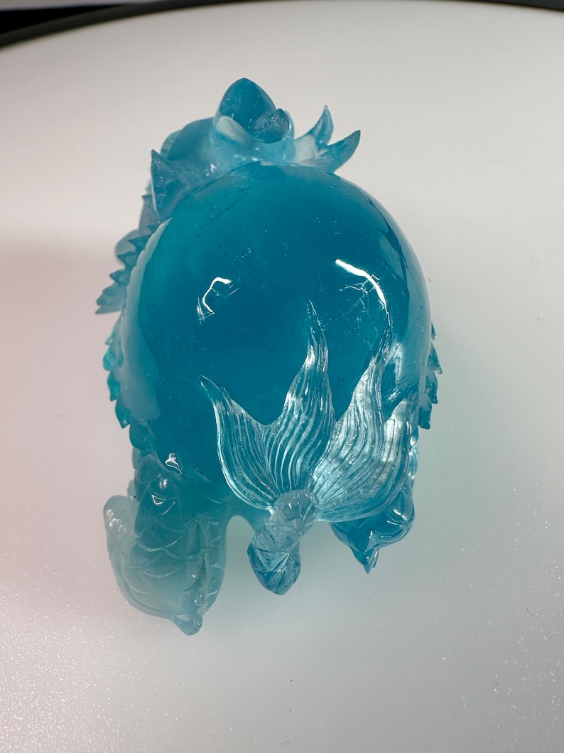 May include: A blue glass sculpture of a dragon with intricate details. The dragon is curled up with its tail wrapped around its body.