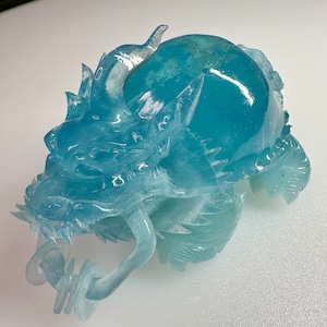 May include: A blue aquamarine dragon sculpture carved from a single piece of stone. The dragon is curled up with its head raised and its mouth open. The sculpture is highly detailed and intricate.