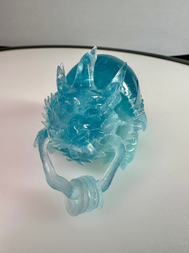 May include: A translucent blue glass dragon sculpture with a curled tail and a detailed face. The dragon is sitting on a white surface.