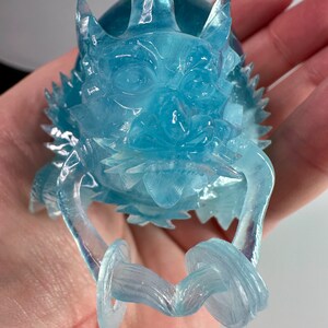 May include: A translucent blue dragon figurine with a detailed face and curled tail. The dragon is made of a clear resin material and has a iridescent finish.