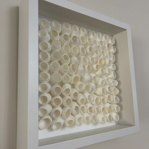 May include: A white framed artwork featuring a grid of white silk cocoons, arranged in a staggered pattern.