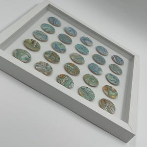 May include: A white framed artwork featuring 28 oval-shaped stones. Each stone has a unique swirling pattern of blue, green, yellow, and brown colors. The stones are arranged in a grid pattern within the frame, creating a decorative piece.