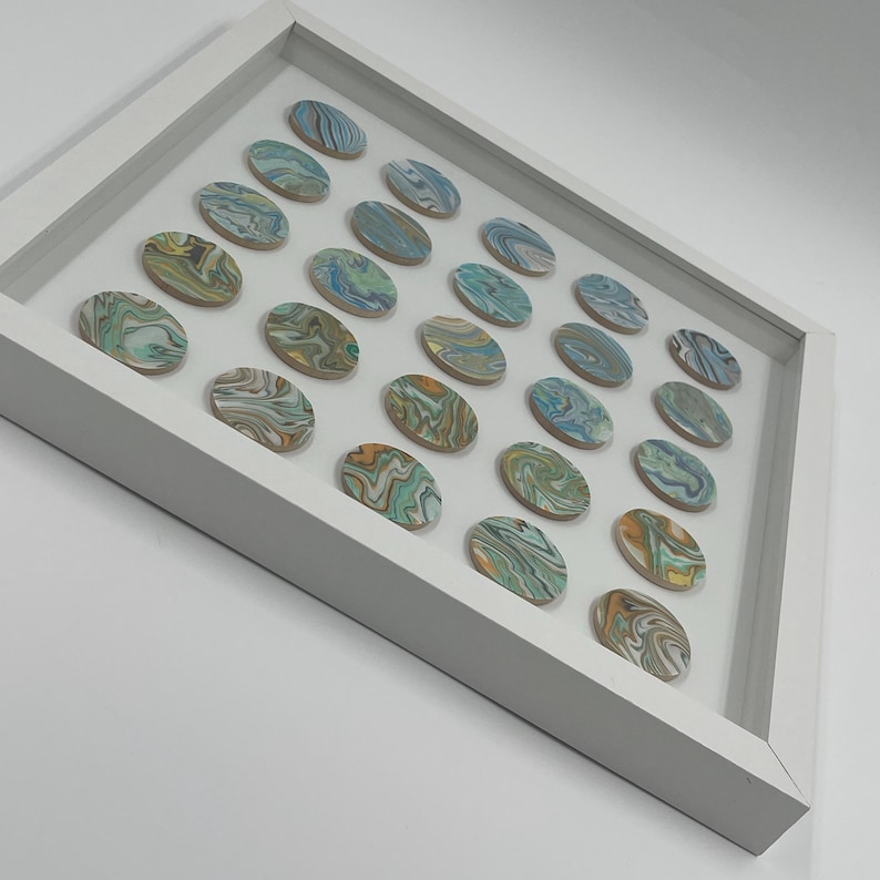 May include: A white framed artwork featuring 28 oval-shaped stones. Each stone has a unique swirling pattern of blue, green, yellow, and brown colors. The stones are arranged in a grid pattern within the frame, creating a decorative piece.
