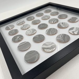 May include: A framed artwork featuring a grid of oval-shaped, gray and white marbled ceramic tiles. The tiles are arranged on a white background within a black frame. The marbled pattern varies across each tile.