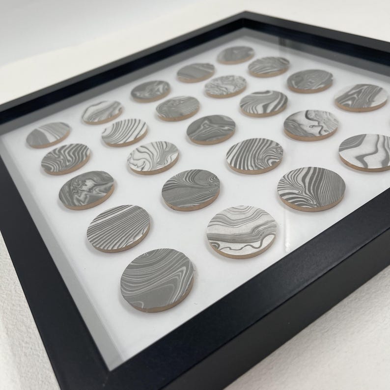 May include: A framed artwork featuring a grid of oval-shaped, gray and white marbled ceramic tiles. The tiles are arranged on a white background within a black frame. The marbled pattern varies across each tile.