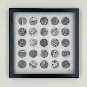 May include: Framed artwork featuring twenty-five circular designs with marbled patterns in shades of gray and white. The artwork is encased in a black frame, creating a modern and minimalist aesthetic. The piece is signed in the bottom right corner.