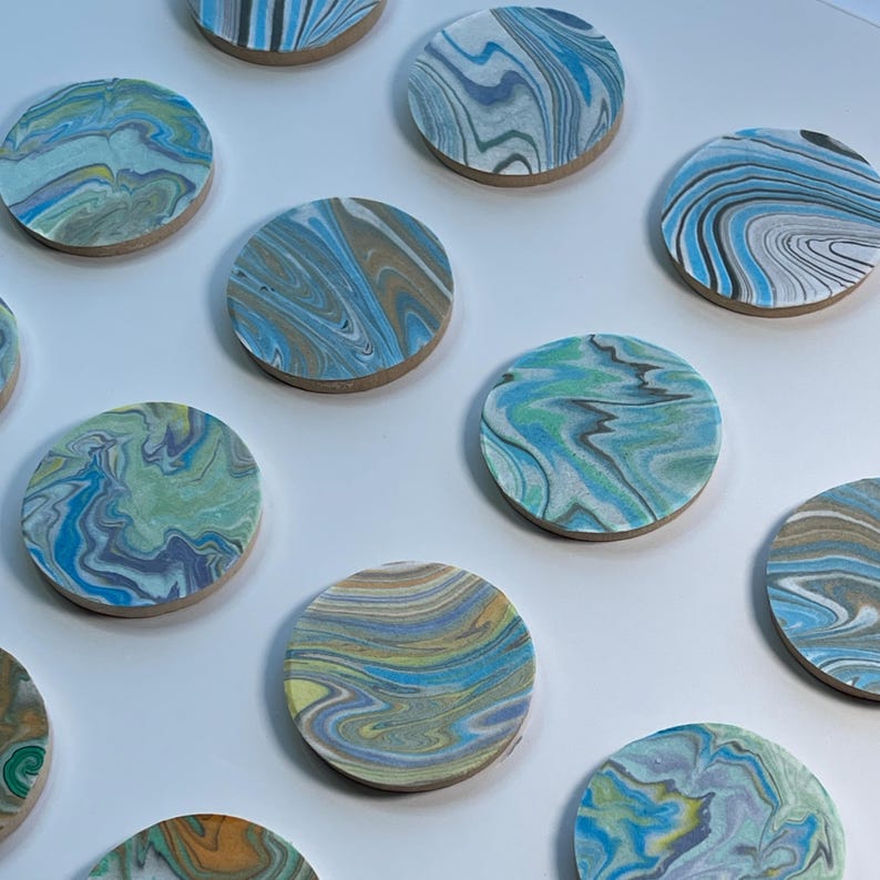 May include: A collection of round coasters with a marbled design. The coasters feature a variety of colors, including blue, green, yellow, and brown, with a swirling pattern. The coasters are made of a light-colored material.