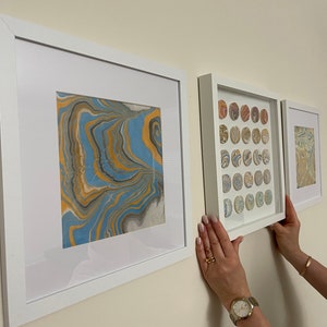 May include: Three framed art pieces with abstract designs in white frames are displayed on a wall. The art features swirling patterns in blue, gold, and gray. The middle frame contains multiple circular designs.