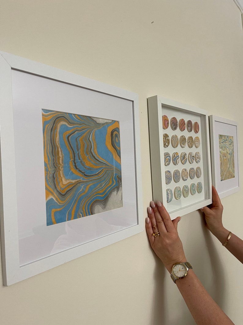 May include: Three framed art pieces with abstract designs in white frames are displayed on a wall. The art features swirling patterns in blue, gold, and gray. The middle frame contains multiple circular designs.