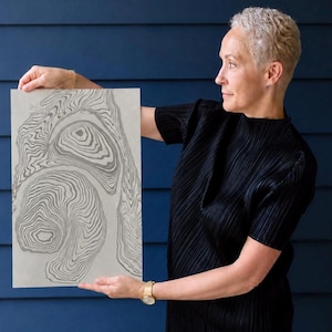 May include: A rectangular art print featuring a light gray background with dark gray, fluid, concentric line patterns that resemble topographic map contours. The design consists of organic, swirling shapes that create a sense of depth and movement across the surface of the paper.